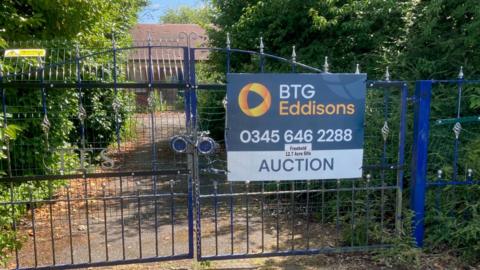 A set of blue metal gates stands at the end of a driveway. Behind them a single-storey building is visible. The gates are locked. On the right hand gate is a sign saying BTG Eddisons Auction.