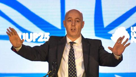 Jon Burrows is speaking in front of a podium at the Ulster Unionist Party Conference in October 2025. He is standing with his arms spread wide, palms facing the audience. He is wearing a dark suit, white shirt and black and white spotted tie.