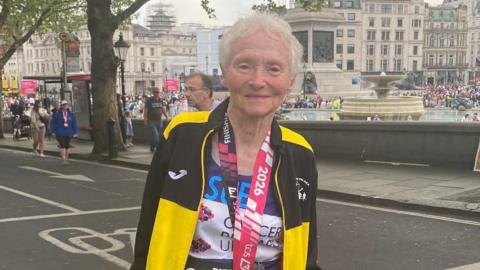 Sue Nicholls is standing on a road in London, wearing a running outfit and a medal. She is smiling.