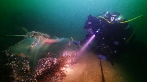 A scuba diver is inspecting a large cauldron embedded into the seabed in the sea. The diver is using a small but powerful light to examine the wreck, and the sea around them is a dark green
