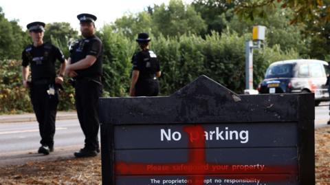 Three police officers in black uniform and hats stand on the pavement next to the hotel sign which reads "No parking". A red St George's cross has been spray painted onto it. Two of them look at the camera, an one has her back turned. There is a black taxi at the side and a speed camera.