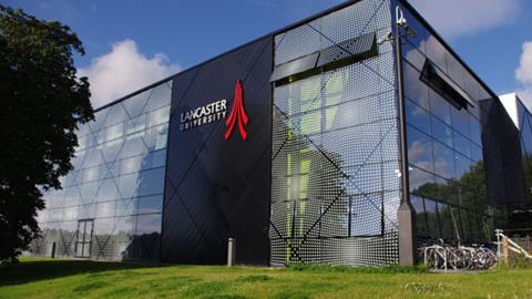 A rectangular glass building at Lancaster University, with a central black column bearing a red 'swoosh' logo and the university name. There is a grass area to the front of the building, and a bicycle rack to the right