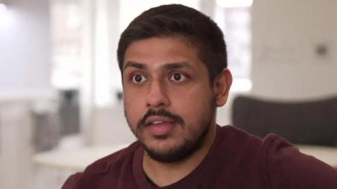 A man with short brown hair, a short brown moustache and beard wearing a maroon T-shirt. He is sitting in a light home