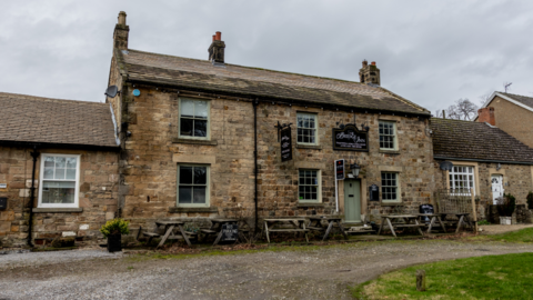 The Bridge Inn is a two-storey stone building with seating area outside. A black sign reading The Bridge Inn hangs above the pale green entrance door.