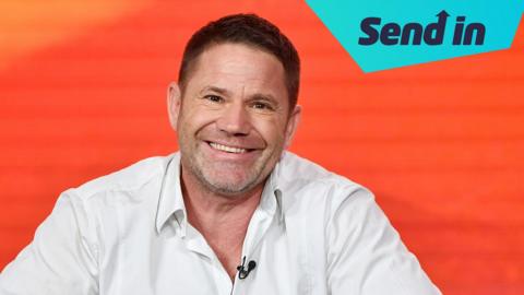 Steve Backshall, a white man with brown hair and grey stubble is smiling with his teeth at the camera. He is wearing a white shirt and the background is a bright orange