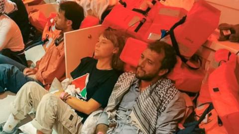 Greta Thunberg is sitting down next tamid a pile of life jackets next to other shipmates. 