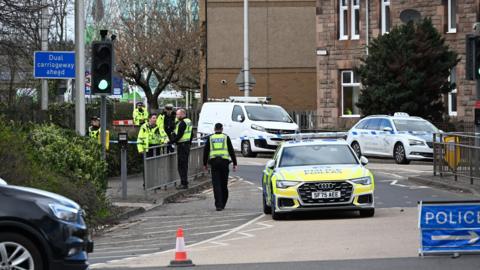 Police officers walk on a pavement. The road has been blocked by a police car and police tape and a cone. A white car and van can be seen behind the police car