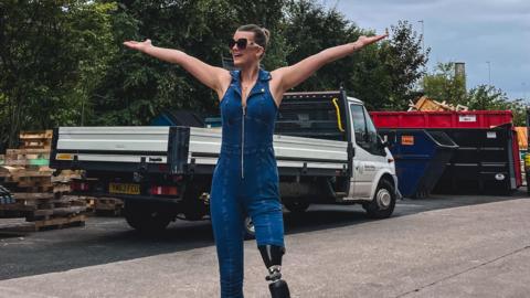 A woman in a denim jumpsuit standing with her arms spread wide at a road side by a lorry and pallets. Her left trouser leg is cut short to reveal a black prosthetic leg. She is wearing sunglasses and has her hair tied back and is smiling at a camera off-centre.