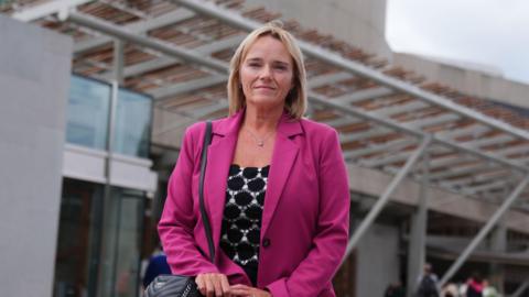 Sandie Peggie wearing a pink jacket and with short blonde hair, outside the entrance to the Scottish Parliament building