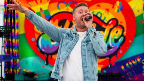 A man in a blue denim jacket and white t-shirt singing, holding a microphone to his mouth. His other hand is stretched out. The backdrop to his performance is a colourful rainbow heart with Pride written on it.
