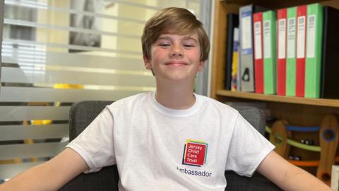 Henry is wearing a white “Jersey Child Care Trust Ambassador” T-shirt sits in a chair with arms resting outward, in an office setting with shelves of folders, books, and small objects in the background.