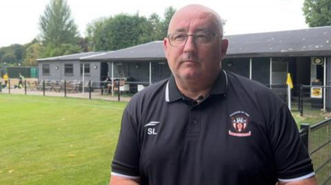 A bald man wearing a black Abbey Hulton United polo shirt stands next to a football pitch with a clubhouse in the background.