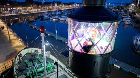 A view from next to the top of a lightship. The light is being cleaned by a man. The rest of the ship is below. Beyond it are boats moored in a marina, as well as buildings lit up at night.