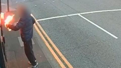 A CCTV grab of Irish Street. To the left of the frame a figure in a blue puffy jacket and dark tracksuit bottoms can be seen holding a flaming object.