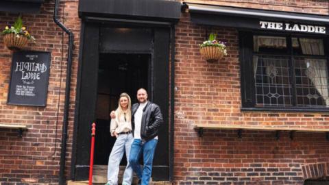 A traditional brick pub exterior with the name ‘The Laddie’ displayed outside. In the doorway there is a man and woman standing close together. Hanging baskets are placed either side of the doorway and a black sign to the left reads ‘Welcome to the Highland Laddie’.
© Ricky Hart