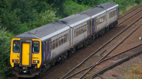 A Northern train on the railway track. Its front is yellow, the sides are grey and purple.