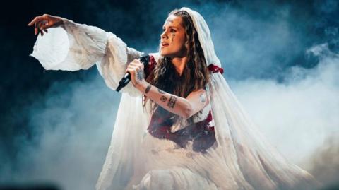 Lelek performing in a white dress with her arm outstretched.