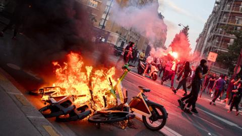 Rioters let off flares while e-bikes burn on a Paris street.