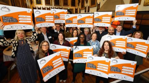 Fifteen people stand in a hall decorated with paintings, while holding up orange and white signs that read: "We commit to challenging racism together for a fairer, stronger North East." They are all looking at the camera and smiling.