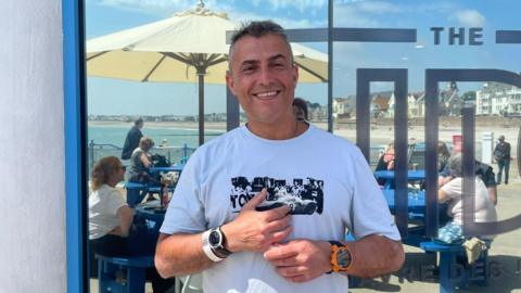 Nelson De Sousa stands in front of a glass window with the partially visible text "THE TID." Behind them is an outdoor seating area with people at blue tables under large umbrellas, and a view of the beach and ocean. He is wearing a white t-shirt with a black graphic design, two watches—one on each wrist—and is holding an object in their hands.
