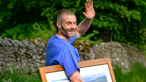 A GP in blue scrubs waves goodbye on a rural road. He is carrying a large framed photo of a rural landscape. 