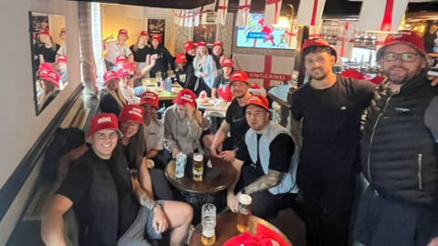Around 20 people wearing red bucket hats sit crammed into a small pub. There are england flags everywhere and a football game is on in the background. 