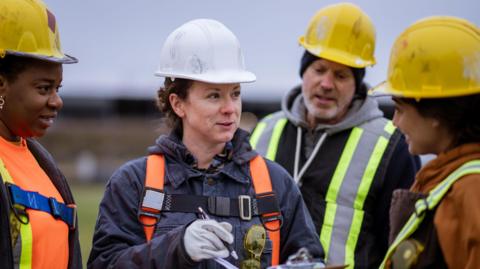 Three women and a man are standing talking to each other. They are all wearing yellow and white hard hats along with high vis jackets 