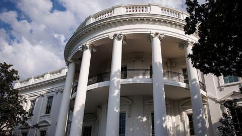 The exterior of the White House in Washtington DC is seen from a low angle and between trees.