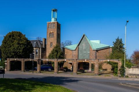 An external view of a church. It is a large brick building with a distinctive green roof and a large cross on the centre of the building.