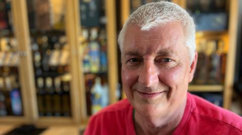 Tom sits in his wheelchair in his living room looking into the camera and smiling. He has short white hair and is wearing a red t-shirt. A wooden display cabinet with a glass front can be seen out of focus behind him. 