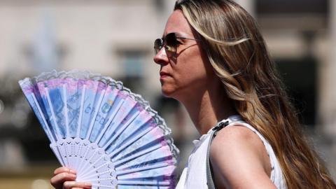 A woman in sunglasses wearing a white top holds a blue and pink fan in London on 24 June 2026.