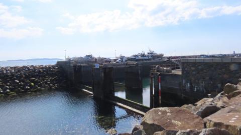 The QEII Marina's gates. Water is low and there is rock armour in the foreground and some boats in the distance.
