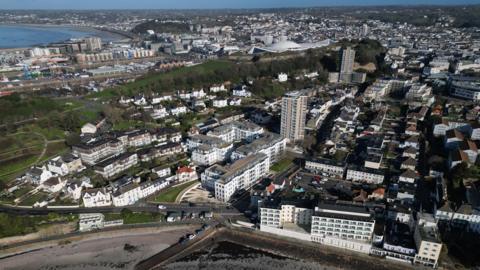 An aerial photo of St Helier. There are a number of large five storey buildings and a few high rise towers. There is also a park visible. There is a bay in the foreground and another in the distance to the left.