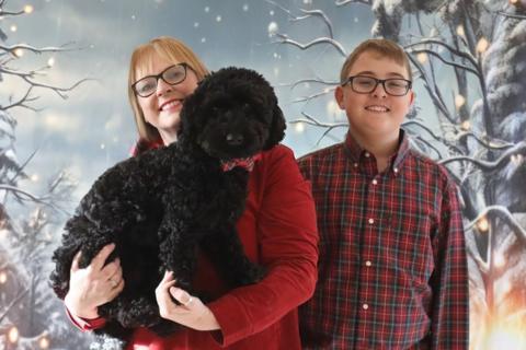 Stefan stands to the right of the image smiling at the camera. He is wearing a checked shirt and dark framed glasses with short brown hair. Sonya stands next to him wearing similar glasses and blond hair. She is wearing a red top and holding a medium size black dog There is a Christmas type background to the photo.
