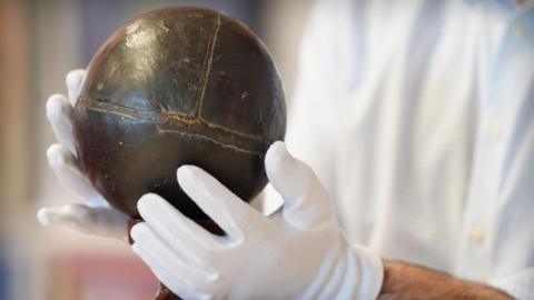 A person wearing white protective gloves holdings a small, shiny leather ball.