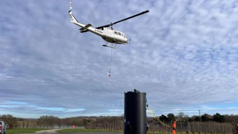 A helicopter picking up the treatment tank to be taken to the Southern Water wastewater treatment works in Pembury. Each tank had to first be picked up from an adjacent orchard, moving to hover above Pembury Wastewater Treatment Works