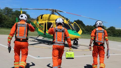 Paramedics and doctors in orange flightsuits walk towards a yellow and green helicopter.