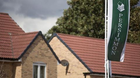 Generic image of new-build houses with red-tiled roofs. There is a pole in the foreground with a green flag bearing the house builder's name and logo.
