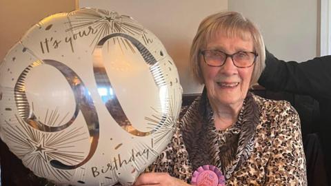 Joan Airey has blonde / grey hair, glasses, patterned top and is smiling. She is sitting on a sofa holding a white helium balloon with 90 Birthday written on it.