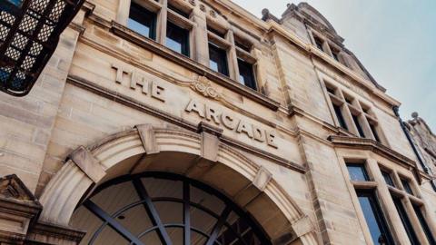 A stone Victorian archway with decorative iron bars inside. Above the archway is The Arcade engraved into the stone masonry. There are windows in the building with stone mullions.