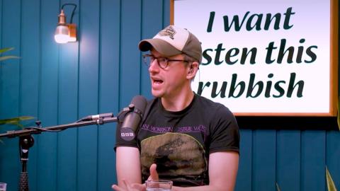 John Robins wears glasses, a dark grey T-shirt and a baseball cap. He speaks into a mic