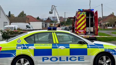 A Leicestershire Police car is parked in front of a cordon as Leicestershire and Fire Rescue Service tackle a fire with an aerial ladder at a bungalow in Colby Drive, in Thurmaston, Leicesteshire. A row of houses, parked cars and a fire service van are visible.