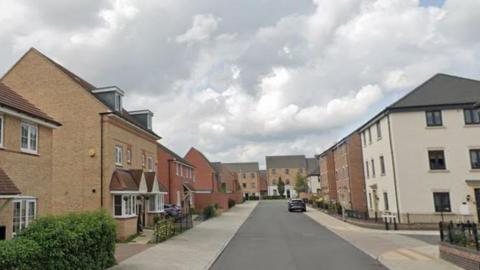 A new housing estate showing two and three-storey brick-built houses around a wide grey road with cream-coloured pavements. A car is parked outside one house. There are dark-coloured railings outside some houses and hedges in some places.