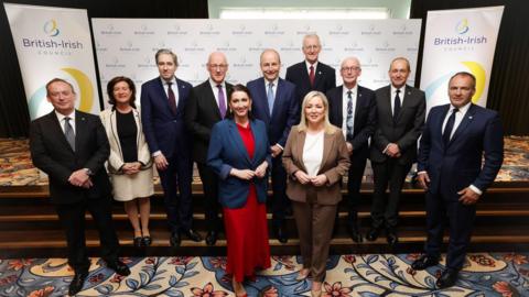 Political leaders from across Ireland, the UK and Crown Dependencies post for a photograph on a set of carpeted steps inside a building during the British-Irish Council summit. Hoardings are behind them with the British-Irish Council logo on.
