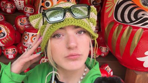 Izabel Rose, a woman with blonde hair and hazel green eyes looks into the camera - she wears a knitted green and beige stripes hat with glasses perched on top and there are Japanese figures in the background