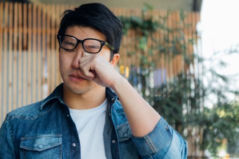 Head shot of young man having allergy symptoms, rubbing his eye with hand.