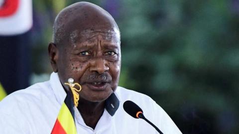 A head and shoulders image of Yoweri Museveni looking at the camera and speaking into a microphone.