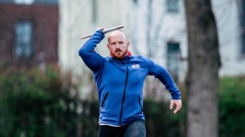 Dan Pembroke, in his run up, holding a javelin wearing a blue GB hoodie