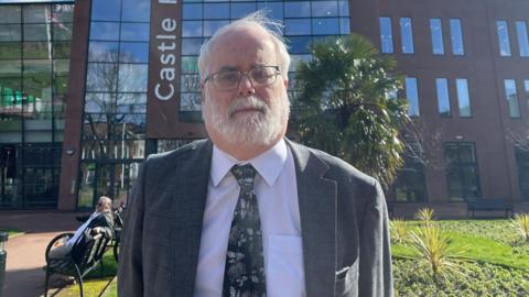 A man with white hair and a white beard, wearing a suit and tie, stands outside the front of a brown building with a park area, trees and people sitting on benches behind him. 