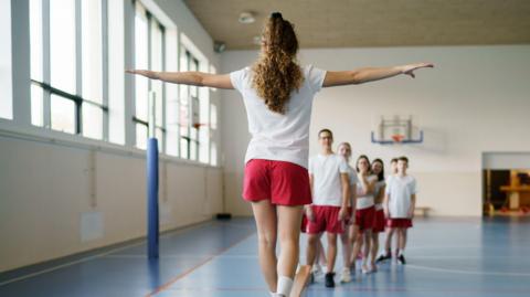Children in PE lesson all wearing red shorts and white top. One girl is balancing on a wooden beam while walking along it with her arms out.